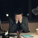 Stressed man holding his head at a desk late at night, illustrating the psychology of procrastination and burnout